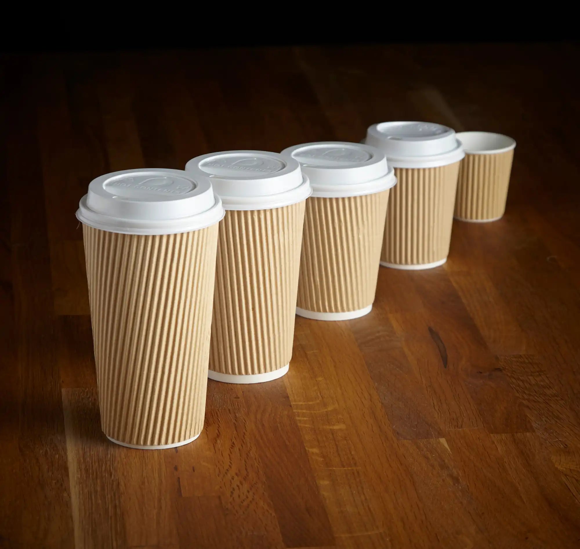 Five disposable coffee cups with ribbed brown sleeves and white lids lined up in a row.